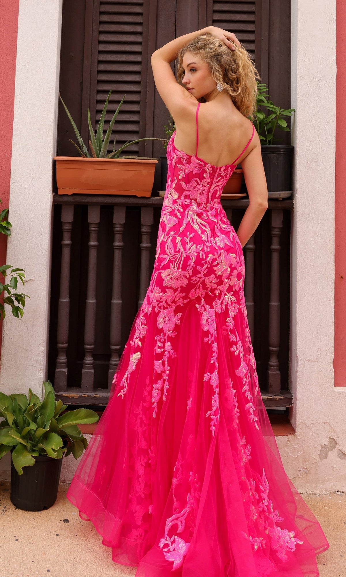 A woman in the Sequin-Lace Long Neon Mermaid Prom Dress Q1390, featuring a bright pink floral design, stands outdoors with her back to the camera and one arm raised, near a window with potted plants.